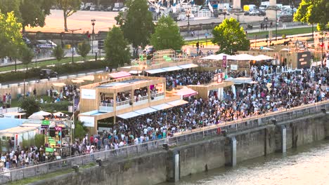 People-crowds-gather-at-Wine-Fair-near-Garonne-river-shore,-Aerial-approach-shot