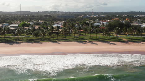 Aerial-view-of-the-beach,-waves-and-a-large-green-area-with-palm-trees-and-the-city-at-background,-Guarajuba,-Bahia,-Brazil