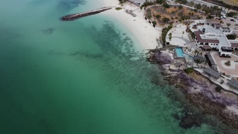 Playa-caimancito-and-la-concha-in-la-paz,-baja-california,-showcasing-the-clear-turquoise-waters-and-white-sandy-beach,-with-coastal-buildings,-aerial-view