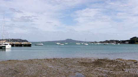 Eine-Handaufnahme-Eines-Strandes-An-Einem-See-Mit-Einigen-Auf-Dem-Wasser-Schwimmenden-Booten-Und-Einem-Vulkan-Im-Hintergrund-An-Einem-Windigen-Tag-Mit-Einigen-Wolken-In-Auckland,-Neuseeland