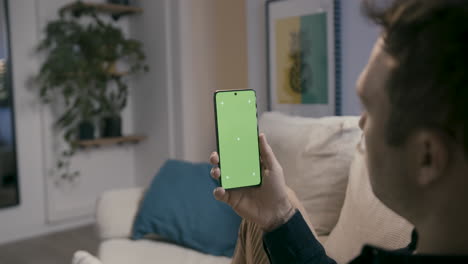 Man-holding-a-smartphone-with-green-screen,-looking-while-sitting-in-the-comfort-of-his-home