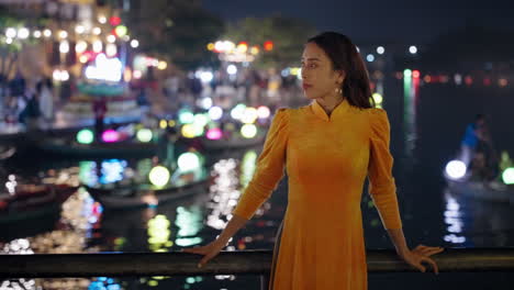 Slow-motion-of-young-Vietnamese-woman-in-traditional-dress-looking-at-night-river-reflections-in-Hoi-An,-Vietnam