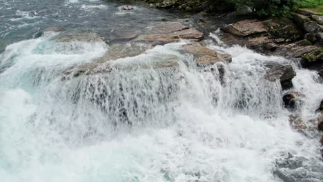 Downwards-tilting-crane-shot-of-crystal-clear-glacial-melt-water-crashing-over-large-rocks-at-the-top-of-a-powerful-waterfall-in-Norway