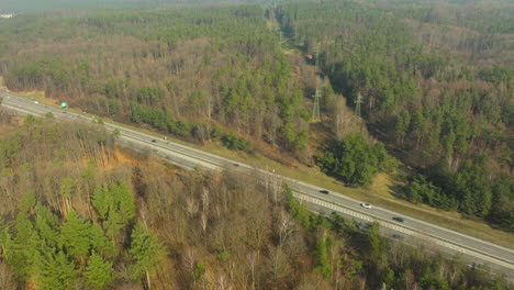 Aerial-shot-capturing-a-highway-slicing-through-a-lush-forest-in-Gdynia-Dąbrowa,-with-vehicles-in-transit-and-power-lines-paralleling-the-road,-illustrating-infrastructure-within-natural-surroundings