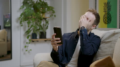 Un-Hombre-Feliz-Y-Sonriente-Usando-Un-Teléfono-Móvil-Mientras-Se-Relaja-En-El-Sofá-De-Casa