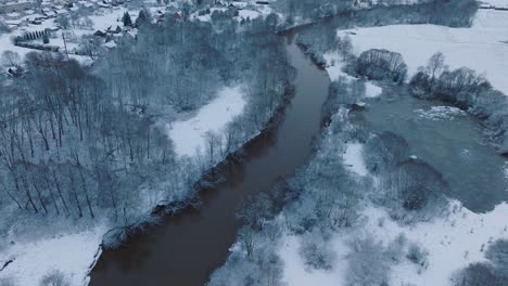 Winding-river-through-snow-covered-forest-in-Abava-river-valley-near-Renda-village-,-aerial-view,-forward-movement,-tilt-up