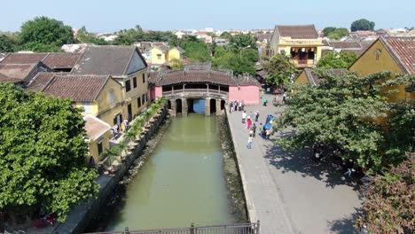 Drone-aerial-view-in-Vietnam-flying-over-Hoi-An-brown-color-river-canal-in-the-city,-small-brick-houses-japanese-pink-old-bridge-on-a-sunny-day