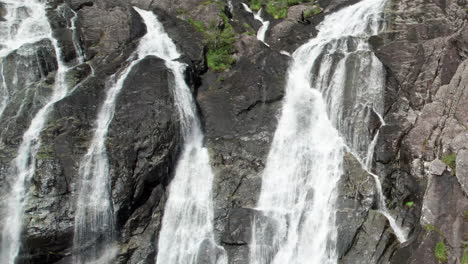 Aerial-dolly-shot-pulling-back-and-descending-from-the-top-of-Laukelandsfossen-Waterfall-in-Norway,-revealing-multiple-gushing-streams-of-water-cascading-over-the-rocky-cliff-face
