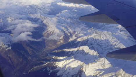 Dramatic-view-out-of-plane-window-of-Swiss-Alps-and-mountains-covered-in-snow-with-blue-sky-and-fluffy-white-clouds