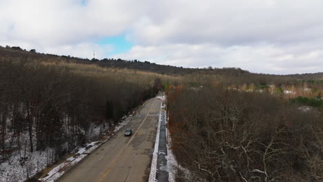 Aerial-rising-shot-of-road-with-car-passing-by-leafless-winter-forest,-Arkansas