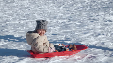 Glücklich-Lächelndes-Kleines-Mädchen-Genießt-Schlittenfahren-Im-Schnee-Bei-Sonnenuntergang,-Gezogen-Von-Mama---Profilansicht-Tracking