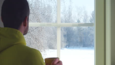 Man-Drinks-hot-tea-or-coffee-looking-at-winter-landscape-through-the-Window,-shifting-focus-concept,-close-up