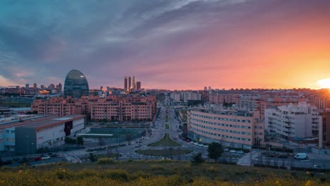 Horizonte-Timelapse-De-La-Moderna-Ciudad-Europea-Madrid-Desde-El-Mirador-De-Las-Tablas-Durante-La-Puesta-De-Sol-Con-Luz-Dorada-Y-Nubes-Rojas-Durante-La-Hora-Azul-Acercar-El-Día-A-La-Noche-Lapso-De-Tiempo-Flores-Amarillas-En-Primer-Plano