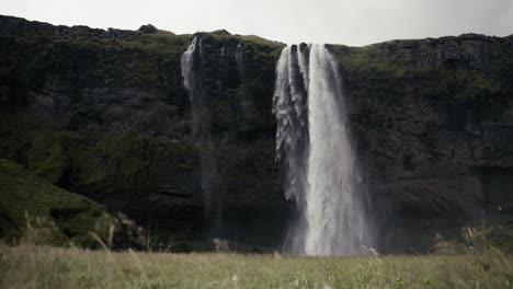 Wide-dolly-shot-of-an-absolutely-stunning-scene-of-a-waterfall,-cliffs,-and-green-field