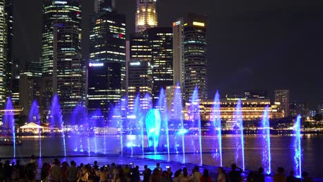 Immersive-SPECTRA-light-and-water-show,-crowd-of-audiences-at-the-Marina-Bay-Sands-event-plaza-with-glass-prism-in-the-center-and-downtown-cityscape-in-the-background,-vibrant-night-of-Singapore-city