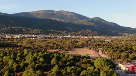 Sinkflug-In-Der-Goldenen-Stunde-Des-Nachmittags-über-Grünen-Kiefernwald-Mit-Gelben-Sommerwiesen-Vor-Dem-Hintergrund-Bewaldeter-Berge-Mit-Sonnigen-Dorfhängen-Und-Blauem-Himmel-In-Avila,-Spanien