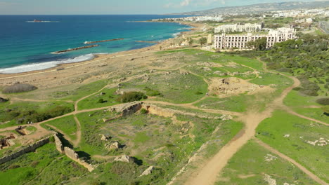 An-aerial-view-of-the-Tombs-of-the-Kings-in-Paphos,-Cyprus,-showing-ancient-ruins-set-against-a-backdrop-of-the-Mediterranean-Sea,-with-modern-buildings-in-the-vicinity