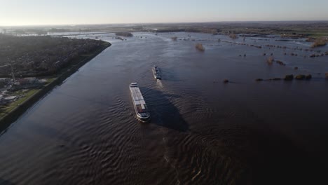 Aerial-following-of-inland-shipping-large-cargo-vessels-leaving-ripple-waves-on-river-IJssel-during-high-water-level-with-flooded-floodplains-of-tower-town-Zutphen-in-The-Netherlands