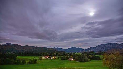 Timelapse-static-shot-of-cloudscape-darkening-over-remote-village-in-Austrian-alps-lying-besides-green-pastures,-forest-and-hills