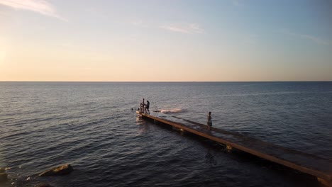 Niño-Salta-Al-Agua-De-Mar-Desde-Un-Largo-Embarcadero-Mientras-Nada-Con-Amigos