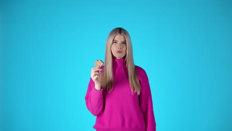 Blonde-woman-with-pink-pullover-takes-a-bit-of-chocolate-cookie,-blue-background