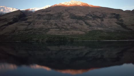 Aerial-view-flying-backwards-to-reveal-perfect-mountain-reflection-at-sunset-with-the-snow-capped-mountain-catching-the-alpenglow-in-Queenstown-New-Zealand