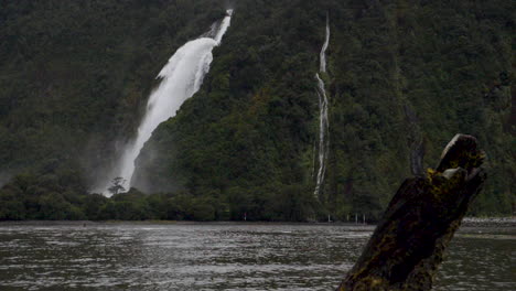 Slow-motion-shot-of-waterfall-in-Milford-Sound-with-a-log-in-the-foreground---Piopiotahi,-New-Zealand