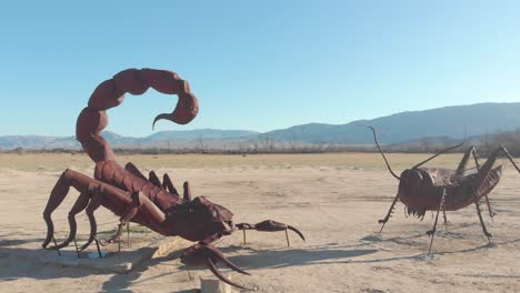 Toma-De-Drones-Semiórbita-De-2-Estatuas-De-Un-Escorpión-Y-Un-Saltamontes-Uno-Frente-Al-Otro-En-El-Desierto-Con-Montañas-Al-Fondo