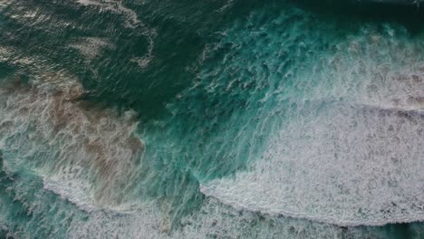 Shot-with-drone-over-sea-waves-in-turquoise-and-white-colors-and-then-rising-towards-the-dark-blue-horizon-and-the-cloudy-sky-letting-the-sun's-rays-pass-into-the-water-in-the-Cantabrian-Sea-Spain