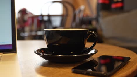 Moviéndose-Hacia-Atrás-Desde-Una-Taza-De-Té-Negra-En-Un-Platillo-Y-Un-Teléfono-Inteligente-Al-Lado-De-La-Mesa-Redonda-De-Madera,-Tiro-Dinámico-Con-Plataforma-Rodante