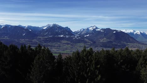 Drone-rise-up-forest-and-reveal-amazing-snow-capped-winter-mountain-landscape-and-cityscape-on-a-beautiful-sunny-day-with-clear-blue-sky-in-Austria