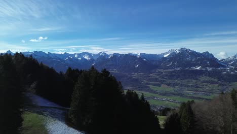 Drohne-Fliegt-Neben-Wald-Und-Enthüllt-Malerische-Winterberglandschaft-Mit-Schneebedeckten-Bergen-Im-Hintergrund-Und-Einer-Kleinen-Stadt-In-Österreich