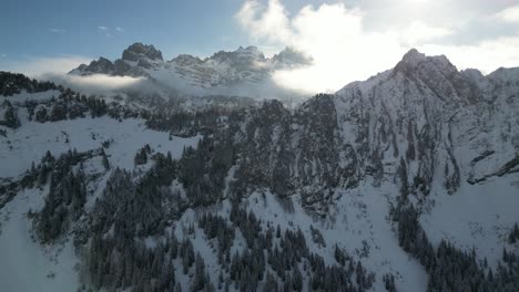 órbita-Paralaje-Alrededor-Del-Bosque-Cubierto-De-Nieve-En-Silla-De-Montar-Con-Crestas-Dentadas-Y-Afiladas-En-El-Fondo-Mientras-Las-Nubes-Difunden-La-Luz-Del-Sol