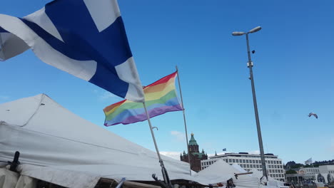 Finnish-National-and-LGBT-Rainbow-Flags-Waving-on-Poles-Above-Shops-in-Market-Square,-Helsinki,-Finland