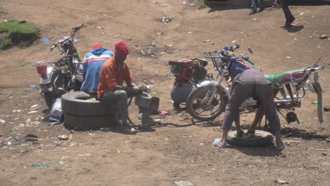 Eine-Lokale-Reparaturwerkstatt-In-Der-Masai-Mara-Mit-Zwei-Arbeitern,-Die-Ein-Motorrad-Im-Sand-Reparieren