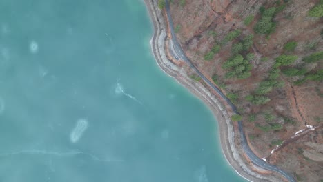 Bird's-eye-view-above-frozen-lake-with-winding-road-along-shoreline-and-base-of-mountain-slopes-in-winter