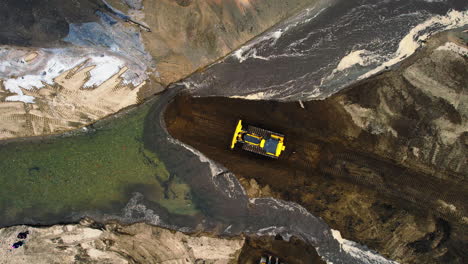 Top-down-aerial-shot-of-yellow-bulldozer-operating-and-moving-soil-in-front-of-river-channel-at-worksite