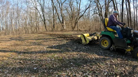 Man-using-John-Deere-utility-tractor-with-attached-rototiller-to-loosen-soil-in-a-wildlife-food-plot-near-a-wooded-area-in-early-spring