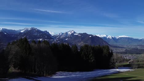 Drone-rise-up-in-sky-fly-above-forest-and-reveal-amazing-winter-mountain-landscape-with-snowcapped-mountains-on-a-sunny-day-in-Vorarlberg-Austria
