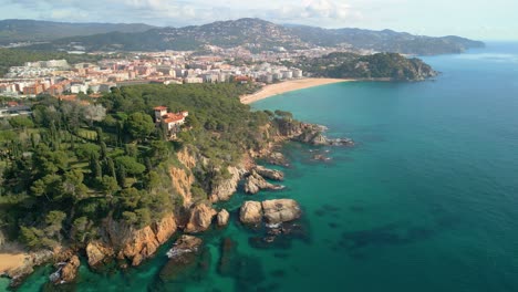 Experience-the-charm-of-Lloret-De-Mar's-aerial-landscape,-featuring-its-azure-sea,-sandy-beaches,-and-exclusive-sites-such-as-Santa-Cristina-and-Cala-Treumal