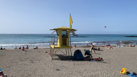 Bustling-beach-day-scene-with-sunbathers,-a-prominent-yellow-lifeguard-tower-stands-watch,-and-a-parasail-drifts-over-calm-sea-waters