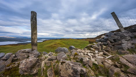 Panorama-motion-timelapse-of-rural-landscape-with-stonewall,-timber-pillars-and-sheep-in-grass-field-and-hills-and-lake-in-distance-during-cloudy-day-viewed-from-Carrowkeel-in-county-Sligo-in-Ireland
