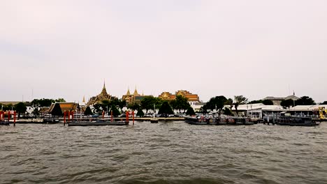 Bangkoks-Lebhafte-Flussszene-Mit-Booten-Und-Dem-Großen-Palast-In-Der-Ferne,-Bewölkter-Tag,-Der-Große-Palast-Im-Hintergrund
