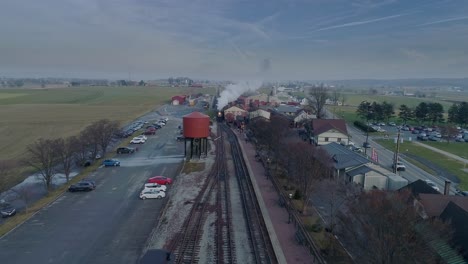 An-Aerial-View-of-a-Train-Station,-with-a-Steam-Passenger-Train,-Pulling-into-the-Station,-Blowing-Smoke,-in-Slow-Motion-on-a-Partially-Sunny-Day
