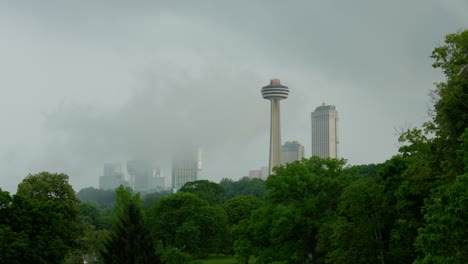 Picturesque-shot-of-the-Skylon-Tower-in-Niagara-Falls,-Ontario,-with-mist-rising-from-The-Falls,-framed-by-a-canopy-of-green,-lush-trees