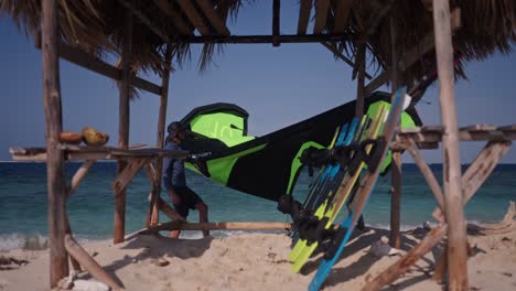 Static-medium-shot-of-a-kiteboarder-holding-a-kite-walking-past-a-small-open-hut-with-boards-leaned-up-inside-and-the-ocean-in-the-background