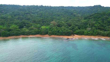 Flying-backwards-over-the-sea-overlooking-the-sundy-beach-with-the-forest-covering-hbd-Sundy-Lodge-Beach,-the-most-expensive-place-at-island,-in-Prince-Island,Africa