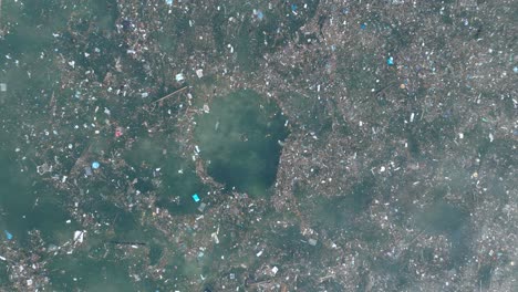 Top-down-quickly-ascending-drone-shot-of-polluted-water-filled-with-trash-floating-over-dead-coral-reef-in-the-turqouise-tropical-water-of-Bali-Indonesia