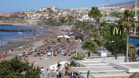 Bustling-Costa-Adeje-beach-in-Tenerife-teems-with-sun-seekers,-thatched-parasols,-sunbathing,-blue-sea-waters,-and-palm-trees-against-a-backdrop-of-resorts-hotels