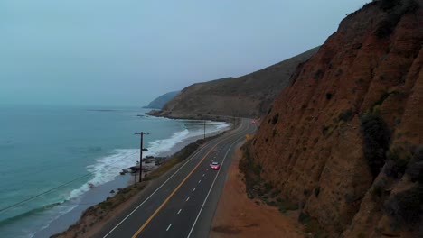 Long-establishing-drone-shot-following-a-car-as-it-pulls-out-onto-Pacific-Highway-One,-near-Malibu-on-a-grey-and-foggy-morning